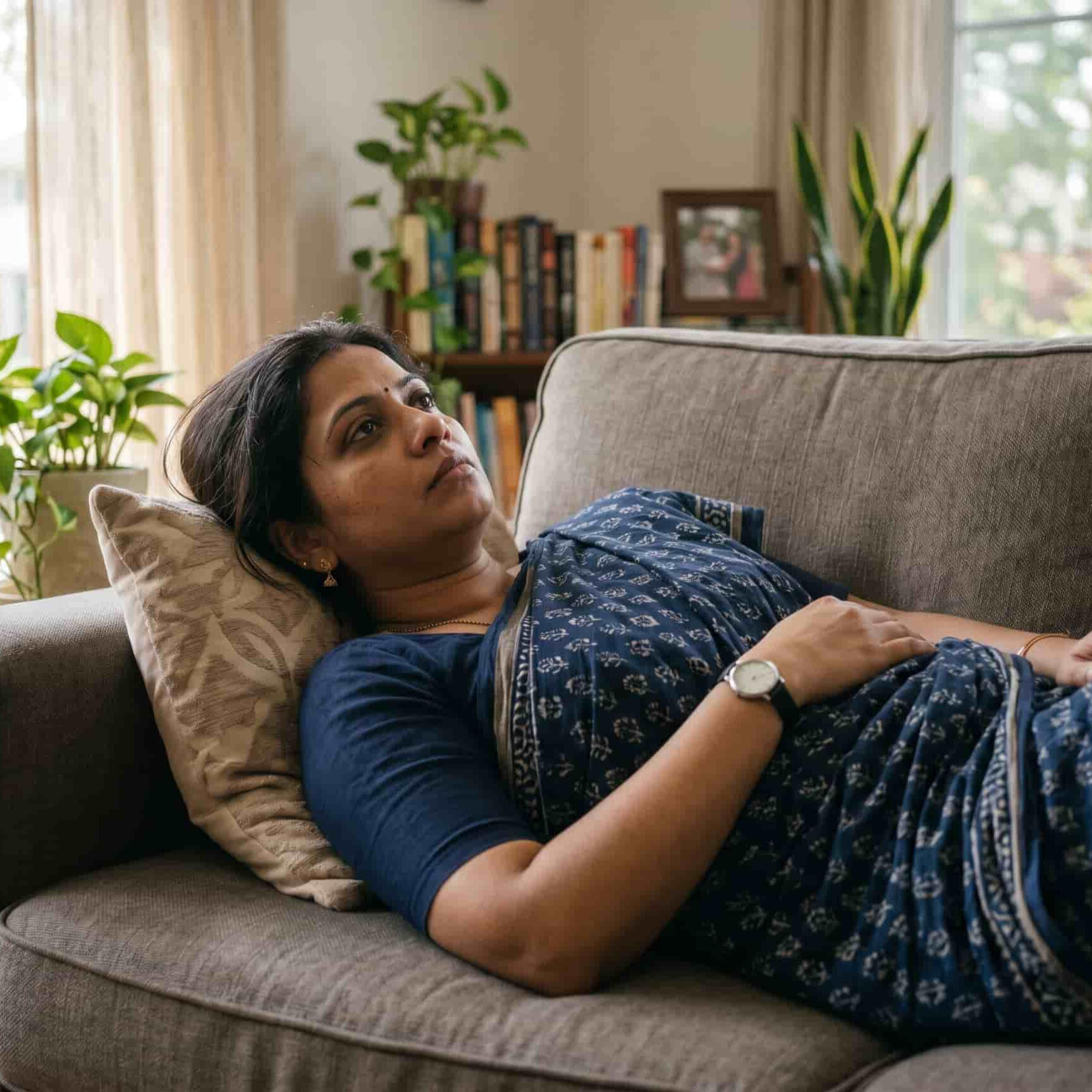 Exhausted Indian woman lying on a living room sofa in a saree. An exhausted Indian woman wearing a blue saree lies on a living room couch looking up at the ceiling, depicting the physical fatigue and emotional burnout of navigating uncoordinated medical care.